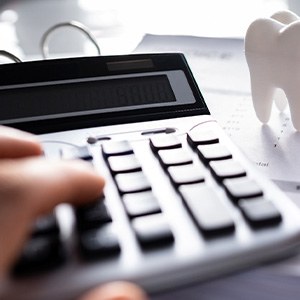 Hands holding pen and touching calculator in front of model tooth and invoices