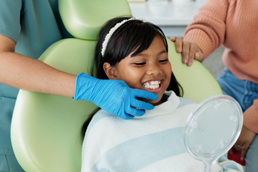 Girl in green dental chair smiling into mirror with dentist touching her chin